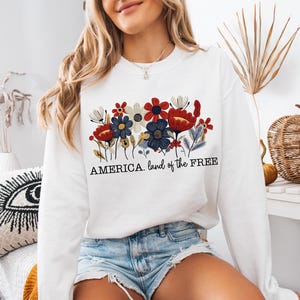 May include: White sweatshirt with a floral design in red, white, and blue. The text "AMERICA. land of the FREE" is printed below the flowers.