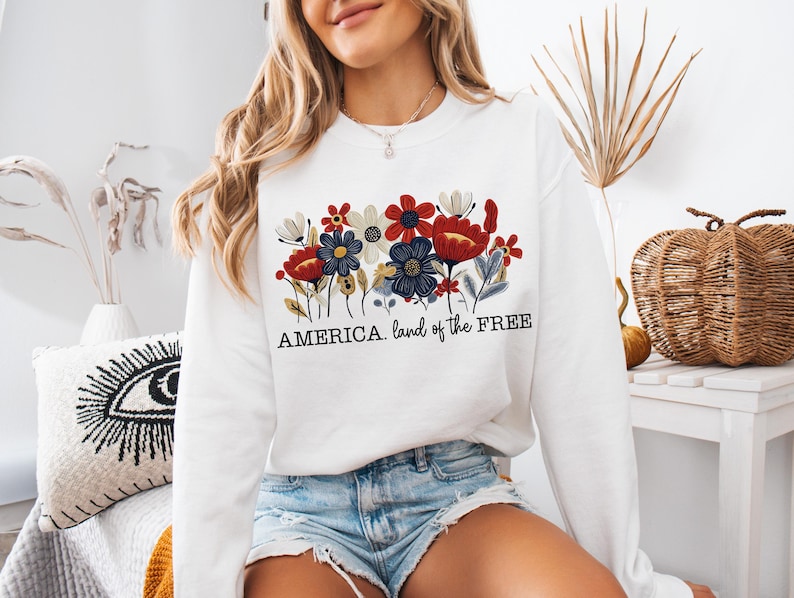 May include: White sweatshirt with a floral design in red, white, and blue. The text "AMERICA. land of the FREE" is printed below the flowers.
