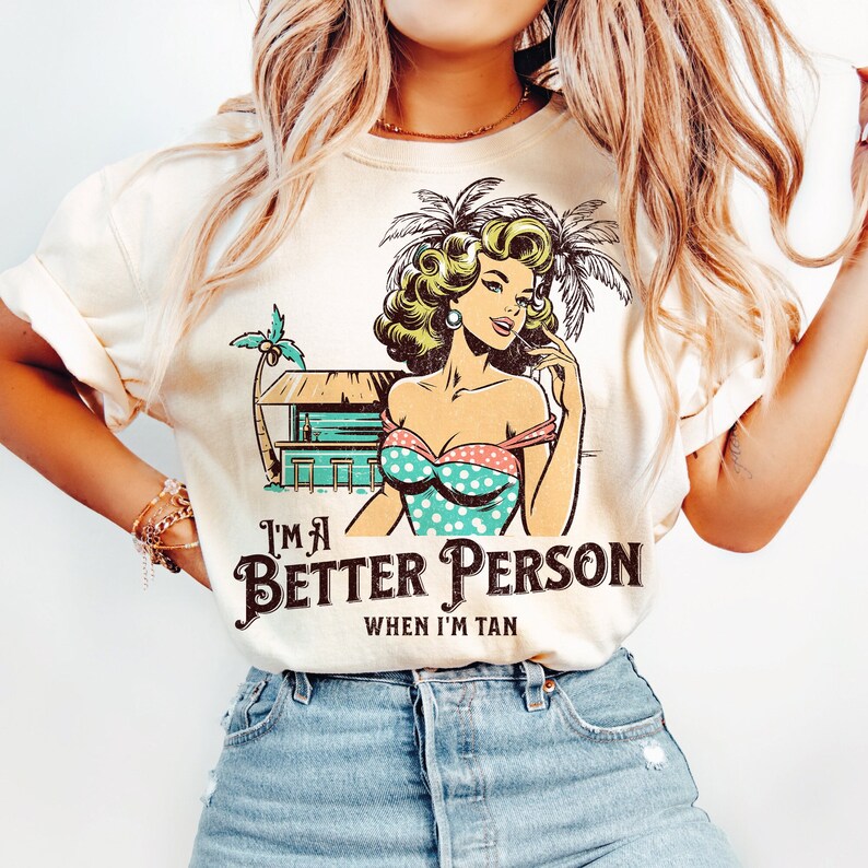 May include: A white t-shirt with a retro-style illustration of a woman in a swimsuit and palm trees. The text "I'm a Better Person When I'm Tan" is printed on the shirt.