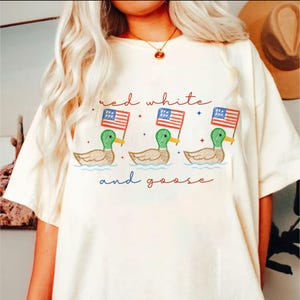 May include: Cream-colored t-shirt featuring a whimsical design of three ducks holding American flags, with the text "red white and goose" in red and blue. The ducks are depicted in green, brown, and yellow.