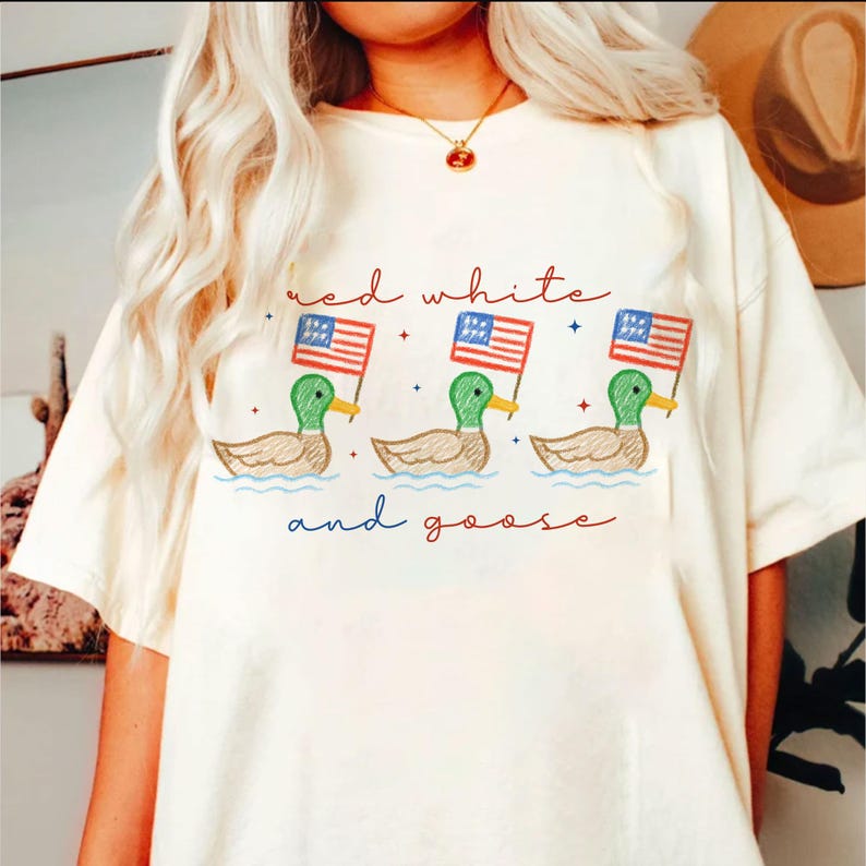 May include: Cream-colored t-shirt featuring a whimsical design of three ducks holding American flags, with the text "red white and goose" in red and blue. The ducks are depicted in green, brown, and yellow.