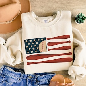 May include: Cream-colored sweatshirt featuring a baseball-themed design. The graphic depicts a baseball and bats arranged to resemble the American flag. The sweatshirt is paired with blue jeans, a hat, and sandals, suggesting a casual, sporty theme.