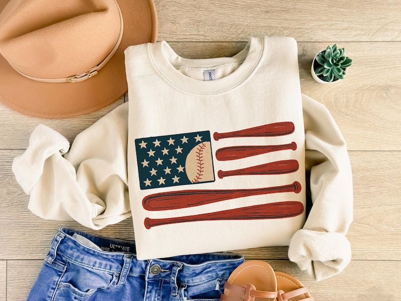 May include: Cream-colored sweatshirt featuring a baseball-themed design. The graphic depicts a baseball and bats arranged to resemble the American flag. The sweatshirt is paired with blue jeans, a hat, and sandals, suggesting a casual, sporty theme.
