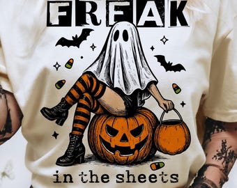 May include: Cream-colored t-shirt with a Halloween-themed graphic. The design features a ghost figure sitting on a carved pumpkin, with the words "FREAK in the sheets" printed above and below the image. Includes bats and candy corn.
