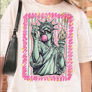 Puede incluir: Camiseta blanca con un gr&aacute;fico de la Estatua de la Libertad con gafas de sol y soplando una burbuja de chicle. La imagen est&aacute; enmarcada con un borde rosa y verde. La Estatua de la Libertad sostiene una antorcha y hace la se&ntilde;al de la paz.