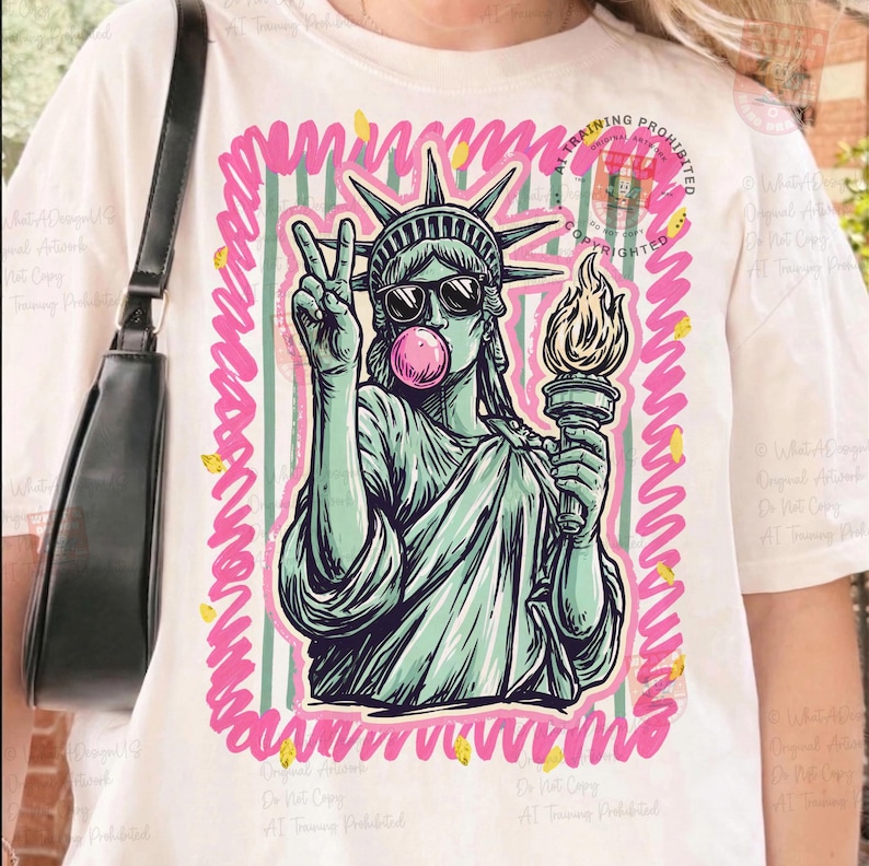 Puede incluir: Camiseta blanca con un gr&aacute;fico de la Estatua de la Libertad con gafas de sol y soplando una burbuja de chicle. La imagen est&aacute; enmarcada con un borde rosa y verde. La Estatua de la Libertad sostiene una antorcha y hace la se&ntilde;al de la paz.