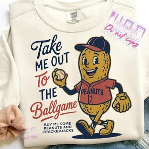 May include: Cream-colored sweatshirt with a baseball-themed graphic. The design features a smiling peanut character in a baseball uniform holding a baseball, with the text "Take Me Out To The Ballgame".