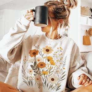 May include: A white sweatshirt with a floral print featuring yellow, orange, and white flowers. The flowers are arranged in a bouquet-like design on the front of the sweatshirt.