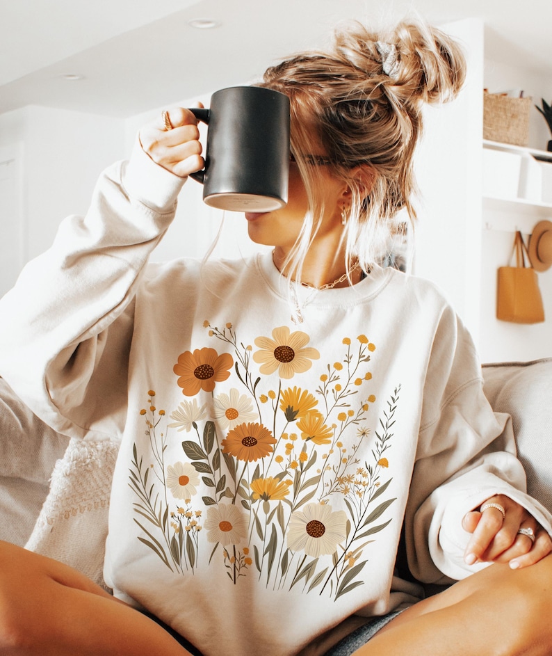 May include: A white sweatshirt with a floral print featuring yellow, orange, and white flowers. The flowers are arranged in a bouquet-like design on the front of the sweatshirt.