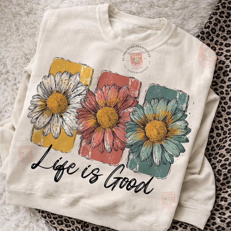 May include: Cream-colored sweatshirt featuring three colorful daisy designs with yellow, pink, and teal petals. Each flower is set against a rectangular backdrop. The phrase "Life is Good" is written in black script below the flowers.