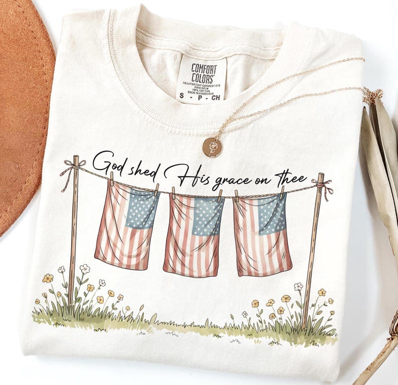 May include: Cream-colored t-shirt with the text "God shed His grace on thee" above a graphic of three American flags hanging on a clothesline. A gold necklace with a pendant rests on the shirt.
