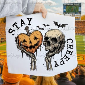 Puede incluir: Gr&aacute;fico blanco de Halloween con una calabaza en forma de coraz&oacute;n y una calavera, ambos sostenidos por manos esquel&eacute;ticas. Las palabras "STAY CREEPY" est&aacute;n impresas en negro. Tambi&eacute;n se incluyen murci&eacute;lagos.