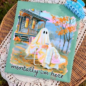 K&ouml;nnte beinhalten: Ein salbeigr&uuml;nes T-Shirt mit einer Grafik eines Geistes, der auf einem Stuhl sitzt und einen Kaffeebecher vor einem Haus mit orangefarbenen Herbstb&auml;umen h&auml;lt. Der Text "mentally i'm here" ist in schwarzer Schrift unter dem Motiv gedruckt.