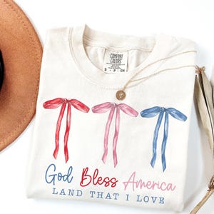 May include: Cream-colored t-shirt with watercolor bows in red, pink, and blue. The text "God Bless America, Land That I Love" is printed below the bows. A gold necklace and a brown hat are also visible.