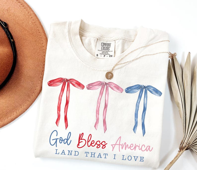 May include: Cream-colored t-shirt with watercolor bows in red, pink, and blue. The text "God Bless America, Land That I Love" is printed below the bows. A gold necklace and a brown hat are also visible.