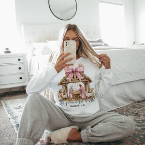 May include: A white sweatshirt with a pink bow and a nativity scene graphic. The text "The greatest gift of all" and "Matthew 1:21" are printed on the sweatshirt.