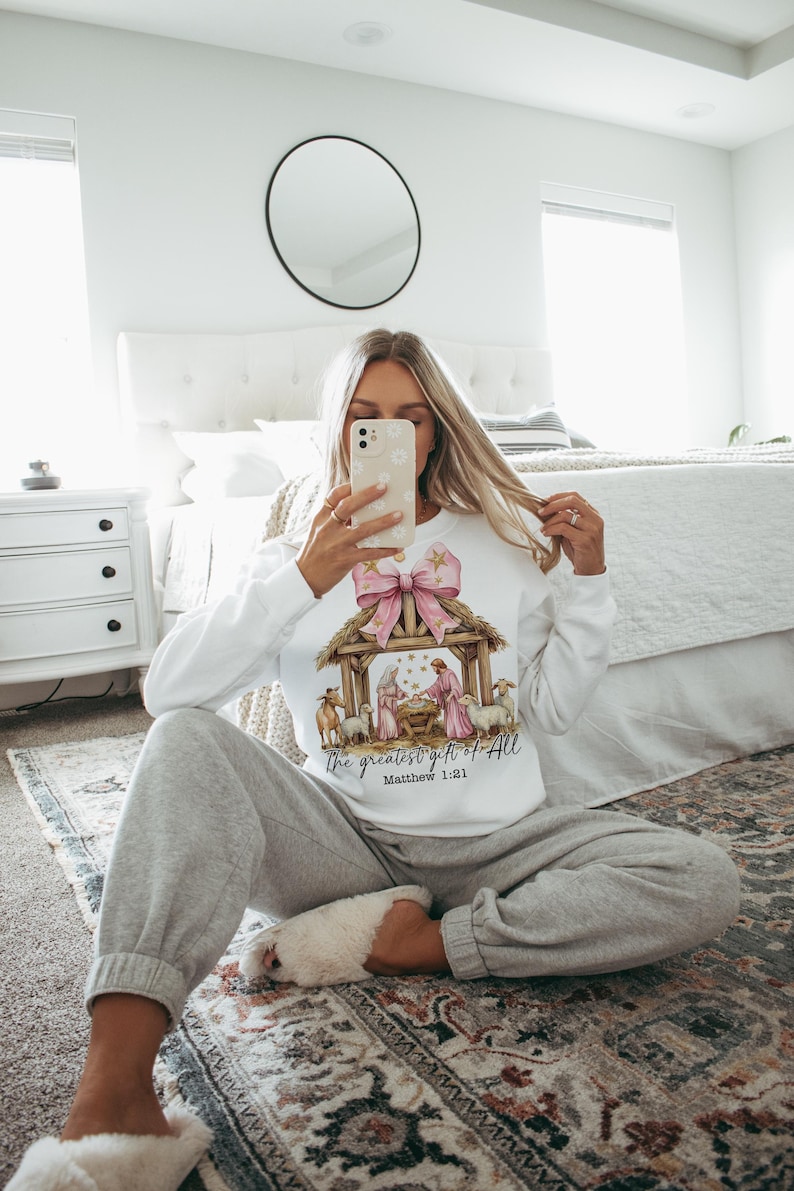 May include: A white sweatshirt with a pink bow and a nativity scene graphic. The text "The greatest gift of all" and "Matthew 1:21" are printed on the sweatshirt.