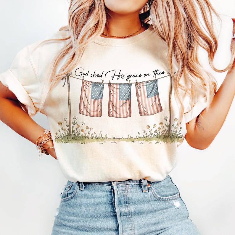 May include: Cream-colored t-shirt featuring a graphic of three American flags hanging on a clothesline, with the text "God shed His grace on thee." The flags have red and white stripes and blue star fields. Floral accents are at the bottom.
