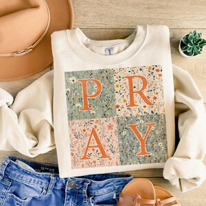 May include: A cream-colored crewneck sweatshirt featuring a square graphic divided into four quadrants. Each quadrant contains a floral pattern in muted tones of green, orange, and pink, with the large orange letters P, R, A, and Y overlaid on top.