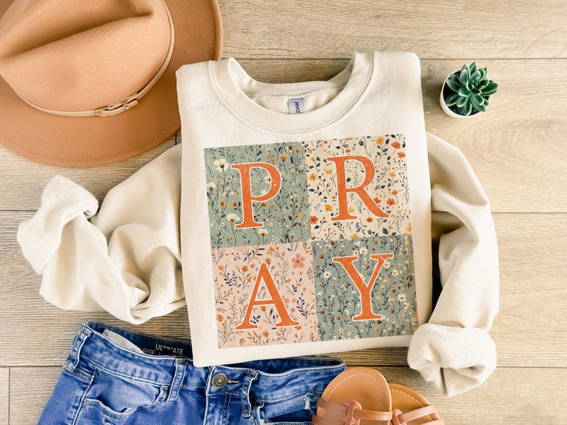 May include: A cream-colored crewneck sweatshirt featuring a square graphic divided into four quadrants. Each quadrant contains a floral pattern in muted tones of green, orange, and pink, with the large orange letters P, R, A, and Y overlaid on top.