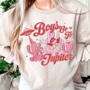K&ouml;nnte beinhalten: Ein beiger Pullover mit einem Grafikdruck eines Cartoon-Cowboys, der ein Einhorn reitet, mit dem Text "Boys Go To Jupiter" in rosa und roten Buchstaben.