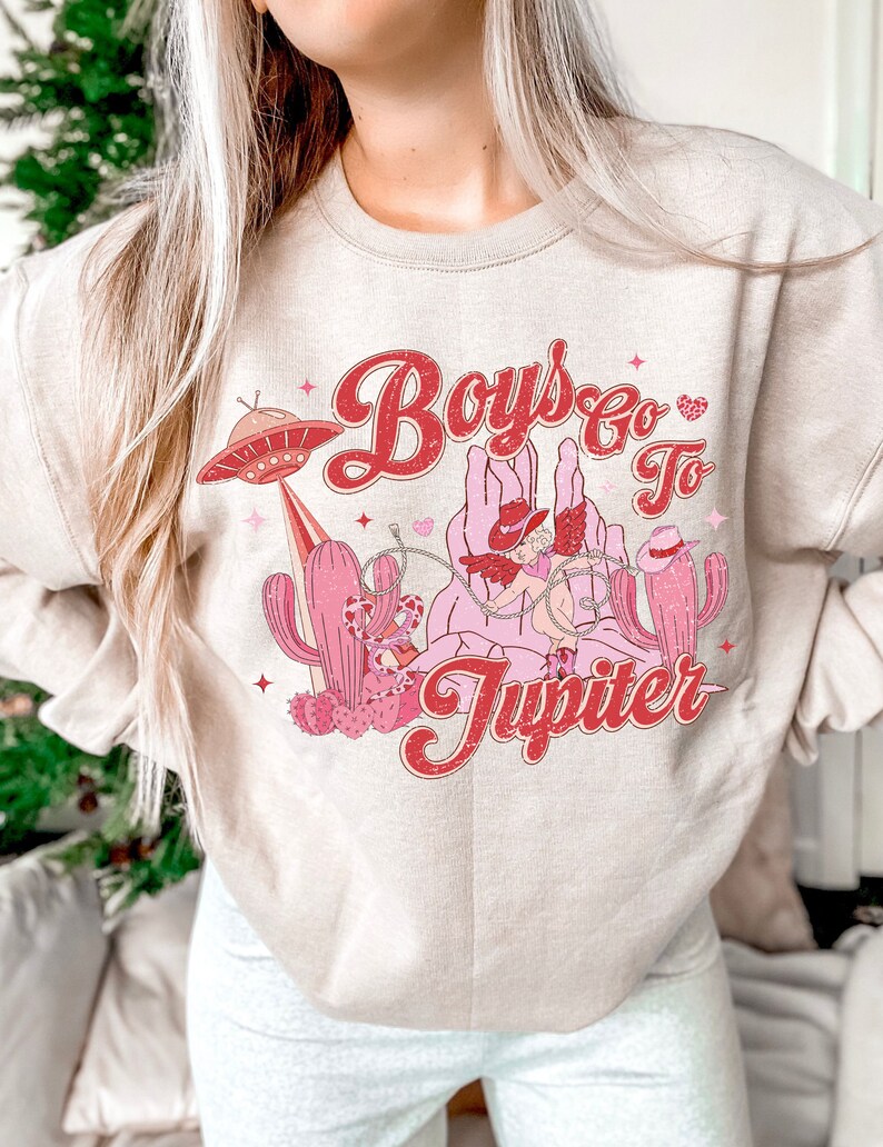 K&ouml;nnte beinhalten: Ein beiger Pullover mit einem Grafikdruck eines Cartoon-Cowboys, der ein Einhorn reitet, mit dem Text "Boys Go To Jupiter" in rosa und roten Buchstaben.