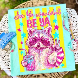 Peut inclure: T-shirt bleu clair avec un raton laveur portant des lunettes de soleil roses et tenant une boisson. Le t-shirt porte l'inscription "See Ya wouldn't BE YA" en rose et jaune, avec des &eacute;toiles.