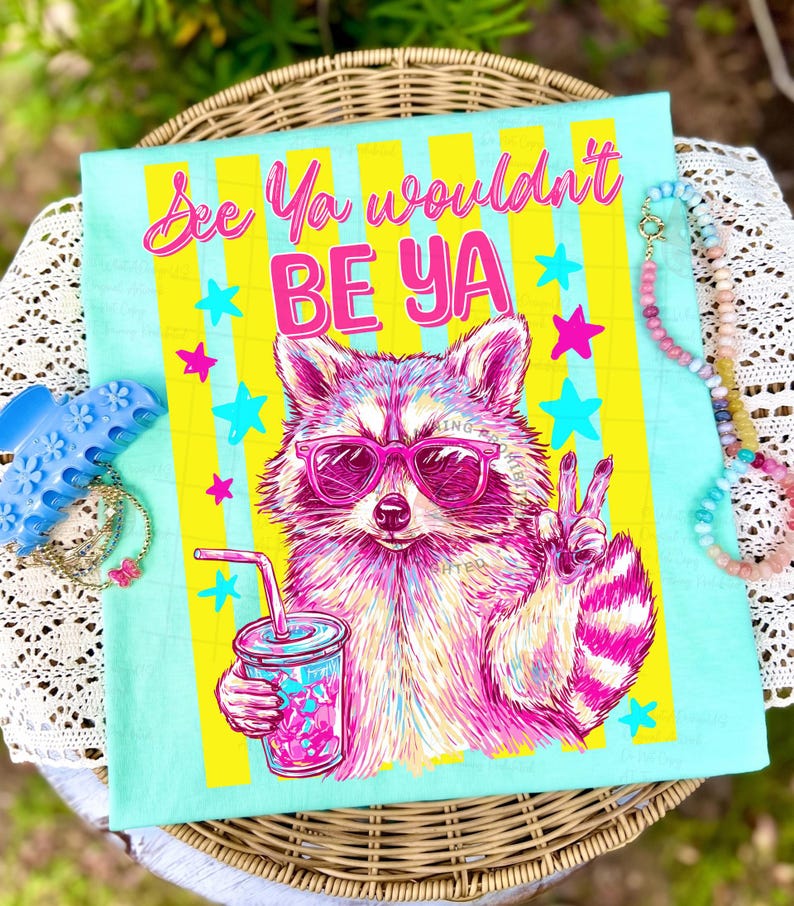 Peut inclure: T-shirt bleu clair avec un raton laveur portant des lunettes de soleil roses et tenant une boisson. Le t-shirt porte l'inscription "See Ya wouldn't BE YA" en rose et jaune, avec des &eacute;toiles.