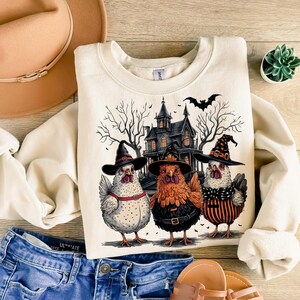K&ouml;nnte beinhalten: Ein cremefarbener Pullover mit einem Halloween-Motiv, das drei H&uuml;hner zeigt, die Hexenh&uuml;te tragen, vor einem gruseligen Haus. Die H&uuml;hner tragen schwarze und orangefarbene Outfits und haben einen skurrilen Look.