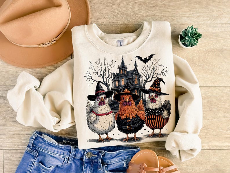 K&ouml;nnte beinhalten: Ein cremefarbener Pullover mit einem Halloween-Motiv, das drei H&uuml;hner zeigt, die Hexenh&uuml;te tragen, vor einem gruseligen Haus. Die H&uuml;hner tragen schwarze und orangefarbene Outfits und haben einen skurrilen Look.