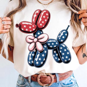 May include: A white t-shirt with a red, white, and blue balloon dog design. The balloon dog has a pink bow and is made of stars and stripes.