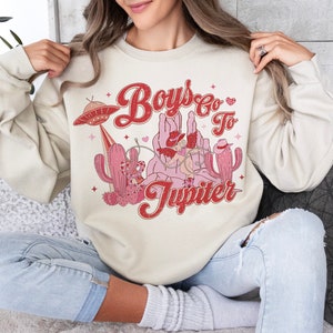 K&ouml;nnte beinhalten: Ein cremefarbener Pullover mit einem Retro-Grafikdesign, das einen Cowboy zeigt, der auf einem Kaktus reitet, mit dem Text "Boys Go To Jupiter" in Rot und Rosa.
