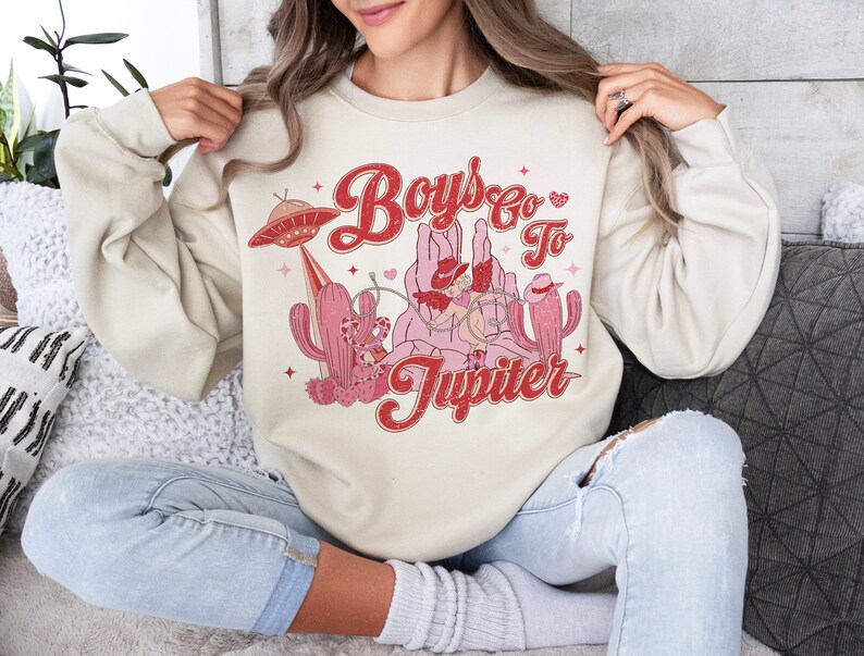 K&ouml;nnte beinhalten: Ein cremefarbener Pullover mit einem Retro-Grafikdesign, das einen Cowboy zeigt, der auf einem Kaktus reitet, mit dem Text "Boys Go To Jupiter" in Rot und Rosa.