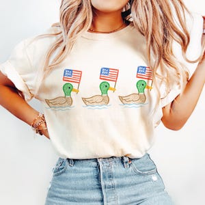 May include: Cream-colored t-shirt featuring a whimsical illustration of three ducks swimming in water, each holding a small American flag. The ducks have green heads, brown bodies, and are set against a blue water background.