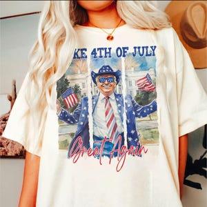 May include: A white t-shirt with a watercolor illustration of a person wearing a cowboy hat and sunglasses holding two American flags. The text "Make 4th of July Great Again" is printed on the shirt.