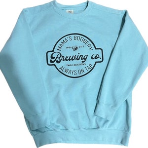 May include: A light blue sweatshirt with a black circular logo that says "Mama's Boobery Brewing Co. Two Locations Always On Tap".