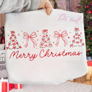 May include: White fabric with a red "Merry Christmas" script. The design includes red bows, Christmas trees, and stars. The fabric is held up by a hand, with a Christmas tree in the background.