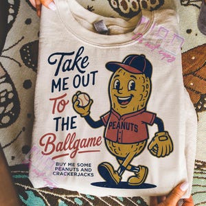 Puede incluir: Sudadera de color crema con un personaje de cacahuete de dibujos animados con uniforme de béisbol, sosteniendo una pelota de béisbol. El texto dice "Take Me Out To The Ballgame" y "Buy Me Some Peanuts and Crackerjacks."
