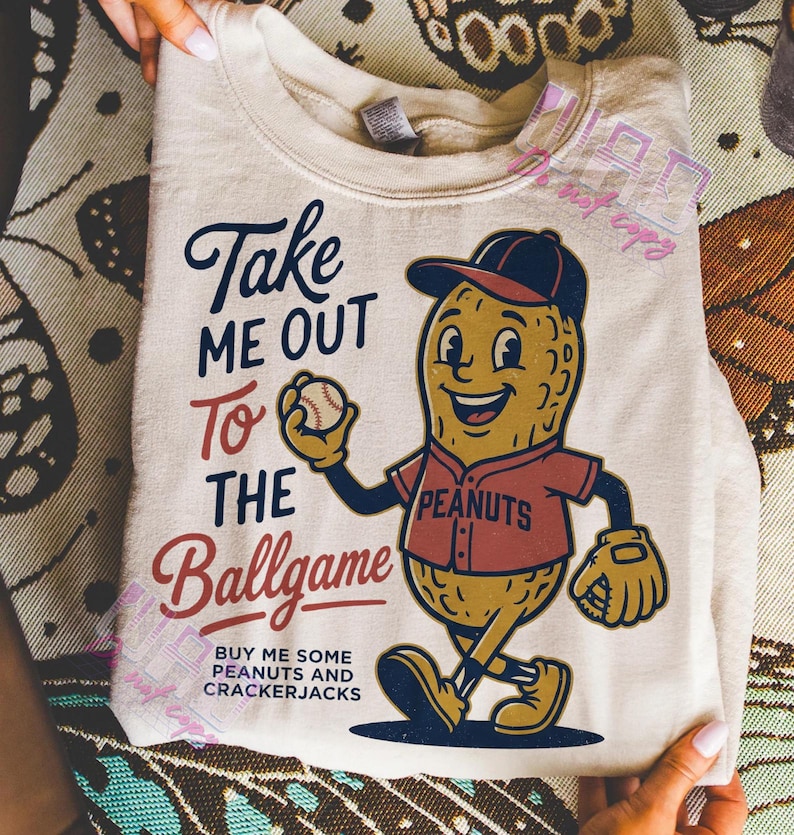 May include: Cream-colored sweatshirt featuring a cartoon peanut character in a baseball uniform, holding a baseball. Text reads "Take Me Out To The Ballgame" and "Buy Me Some Peanuts and Crackerjacks."
