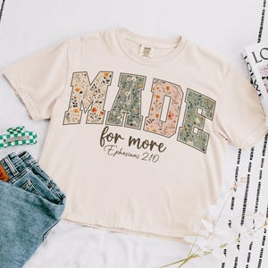 May include: Cream-colored crop top with the words "MADE for more" in floral print. The text "Ephesians 2:10" is below. A pair of blue jeans, a green and white hair clip, and magazines are also in the image.