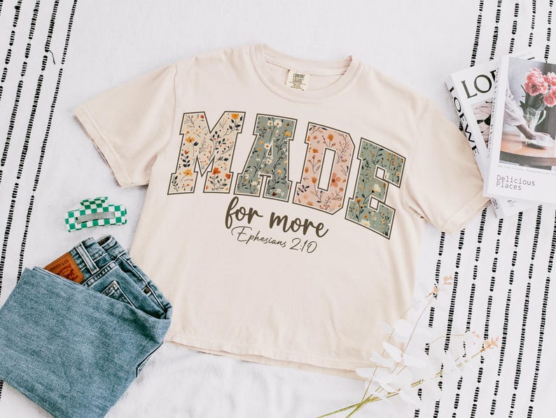 May include: Cream-colored crop top with the words "MADE for more" in floral print. The text "Ephesians 2:10" is below. A pair of blue jeans, a green and white hair clip, and magazines are also in the image.
