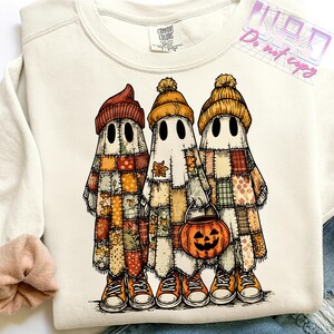 May include: Cream-colored sweatshirt featuring a graphic of three ghost figures in patchwork outfits and orange beanies. The ghosts are wearing orange sneakers and holding a jack-o'-lantern bucket. The sweatshirt has the Comfort Colors brand label.