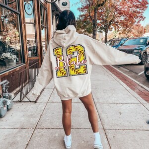 May include: A white hoodie with a yellow softball design on the back. The softball has red stitching and the number 12 with the name "Blake" written in pink.