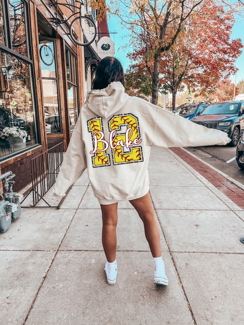 May include: A white hoodie with a yellow softball design on the back. The softball has red stitching and the number 12 with the name "Blake" written in pink.