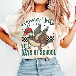 May include: A white t-shirt with a green duck wearing a red hat and a backpack. The text "flying into 100 days of school" is printed on the shirt.