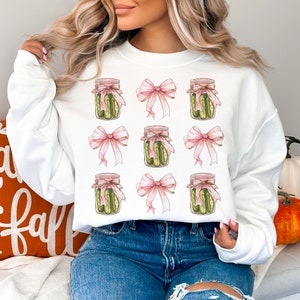 Op de afbeelding: Wit sweatshirt met een herhalend patroon van augurkenpotten gebonden met roze strikken. Het sweatshirt wordt gedragen door een persoon met lang blond haar.