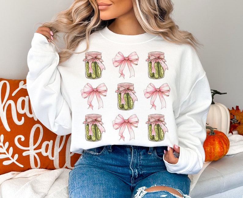Op de afbeelding: Wit sweatshirt met een herhalend patroon van augurkenpotten gebonden met roze strikken. Het sweatshirt wordt gedragen door een persoon met lang blond haar.
