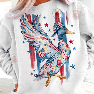May include: White sweatshirt with a patriotic duck illustration. The duck is decorated with American flag patterns, stars, and floral details. Red and blue brushstroke stripes and stars are in the background. The duck wears a matching hat.