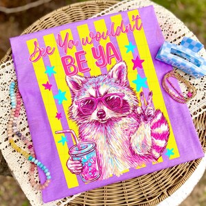 Peut inclure: T-shirt violet avec l'inscription "See Ya wouldn't BE YA" et un raton laveur portant des lunettes de soleil roses, tenant une boisson. Le motif comprend des rayures jaunes et des &eacute;toiles color&eacute;es. Une pince &agrave; cheveux &agrave; carreaux bleus et blancs et un collier de perles sont &eacute;galement visibles.