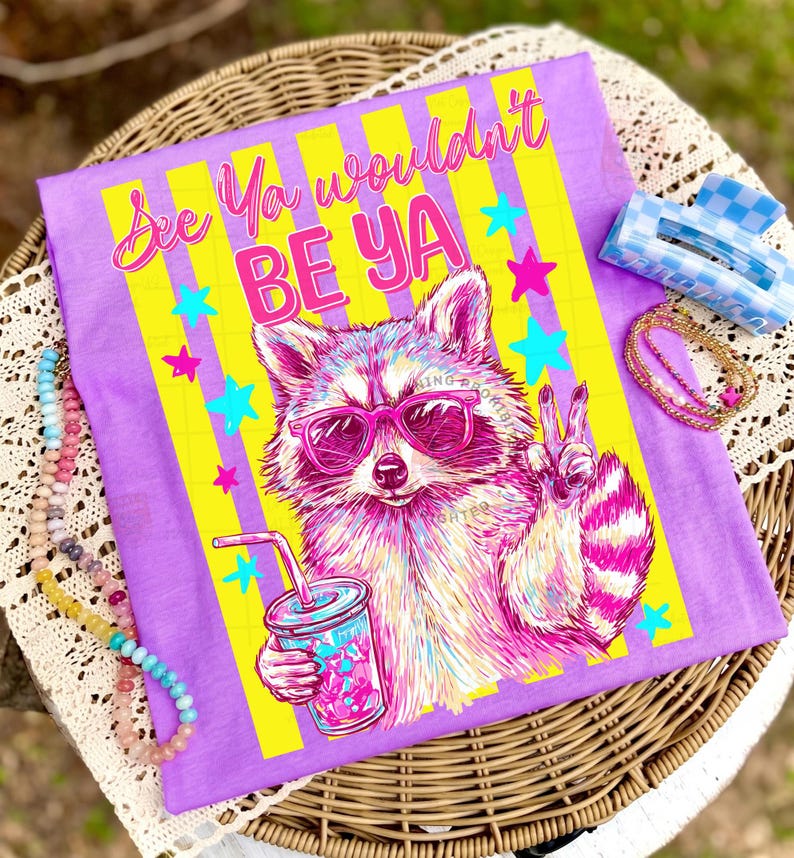 Peut inclure: T-shirt violet avec l'inscription "See Ya wouldn't BE YA" et un raton laveur portant des lunettes de soleil roses, tenant une boisson. Le motif comprend des rayures jaunes et des &eacute;toiles color&eacute;es. Une pince &agrave; cheveux &agrave; carreaux bleus et blancs et un collier de perles sont &eacute;galement visibles.